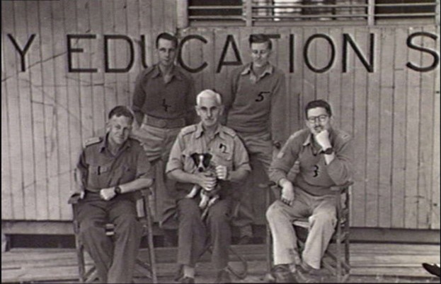 A group of men outside an Education Office. One man is holding a small dog.